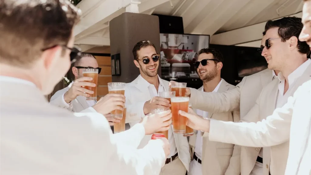 Guys in beige suits and white shirt, that begin a bachelor party in Thailand by cheering with beers.