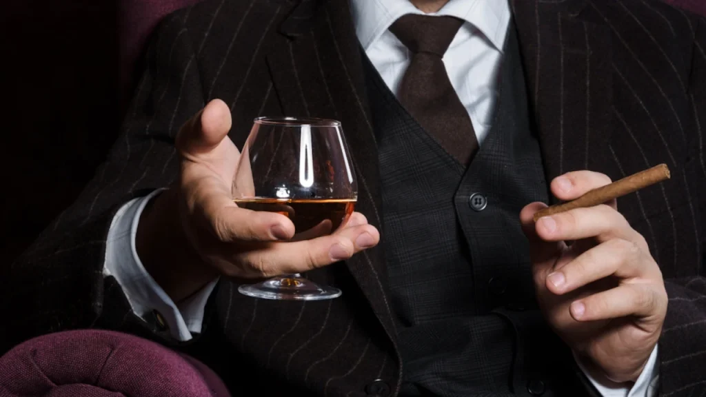 man in a suit smoking a cigar and holding a glass of whisky